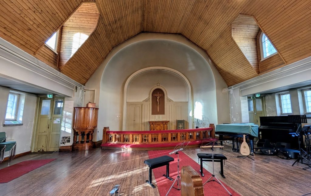 Yleiskuva kirkkosalista. Korkea puinen katto, punaisia mattoja lattialla, saarnastuoli ja alttari, jonka takana krusifiksi.