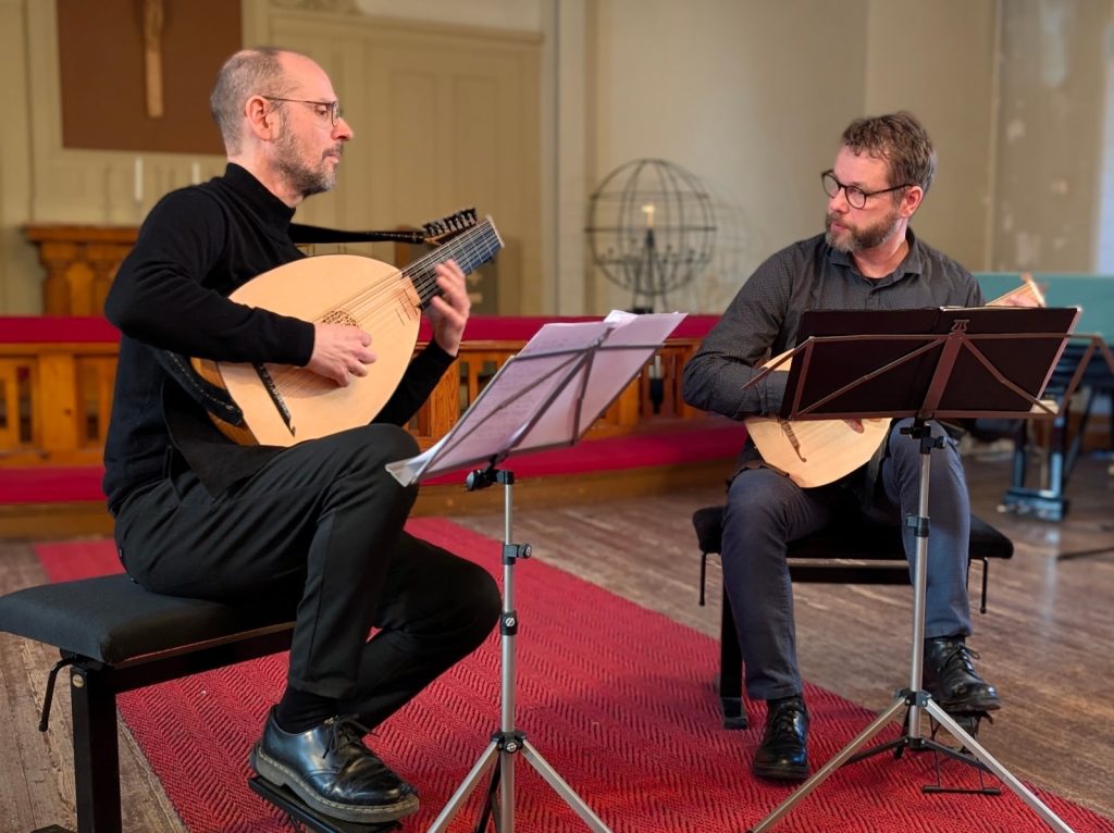 Kaksi tummiin pukeutunutta miestä soittamassa luutuilla yhdessä.