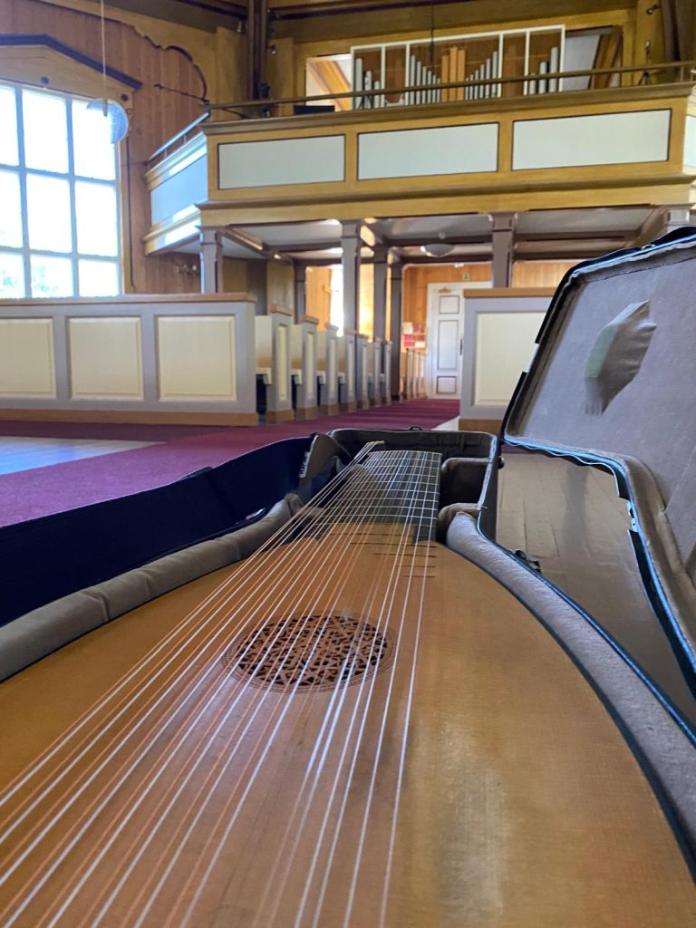 Luuttu lähikuvassa kirkon penkkien ja urkuparven edessä. Soitin on kotelossaan, ja sen kielet näkyvät selvästi etualalla. Taustalla näkyy kirkon vaaleita puupenkkejä, iso ikkuna ja urut parvella. Tiloissa on lämmin, puisista pinnoista koostuva sisustus ja pehmeä matto lattialla.