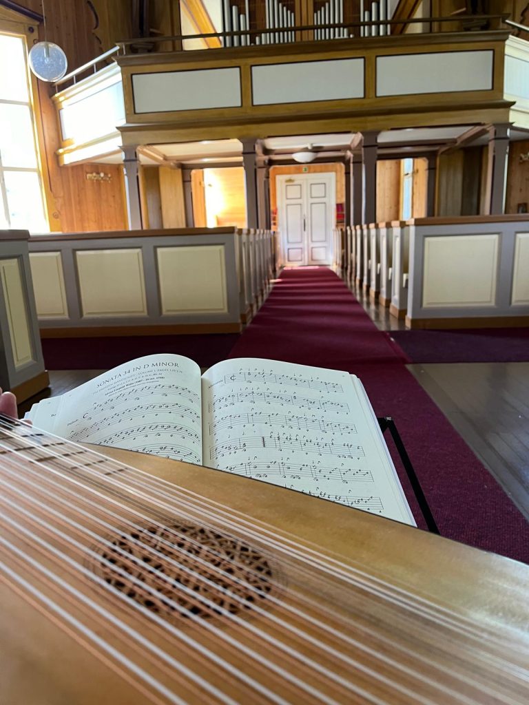 Kuva kirkon sisätilasta. Kuvan etualalalla on luuttu, jonka kielet ja koristeellinen ääniaukko näkyvät selvästi. Luutun takana on nuottikirja, joka on avattu, ja siinä lukee ylhäällä "Sonata 34 in D minor", nuotit ovat käsinkirjoitettua tabulatuuria. Taustalla näkyy kirkon vaaleita penkkejä ja käytävä, jossa on punainen matto. Kauempana edessä on valkoinen ovi ja parvella urkupilitt. Kirkkoon tulee luonnonvaloa vasemmalta ikkunasta.