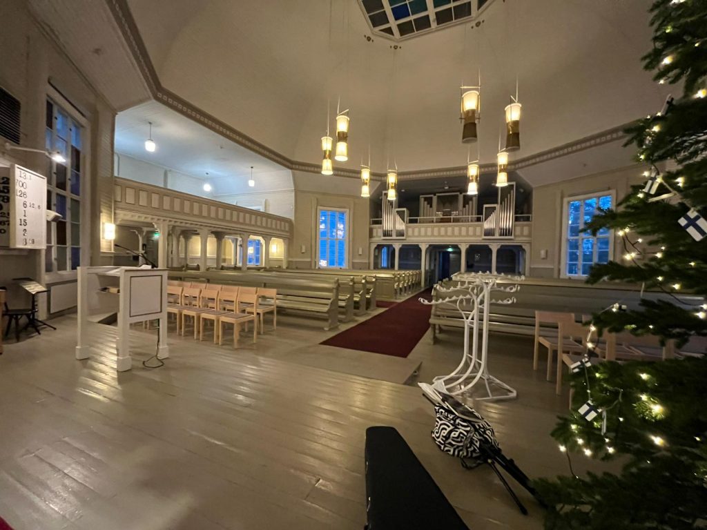 Kirkon sisätila. Kuvassa näkyy vaalea puukirkko, jossa on paljon penkkirivejä, vaalea puiset lattiat, korkea kupolikatto ja riippuvalaisimia. Keskellä kirkkoa kulkee punainen matto alttarille päin. Vasemmalla on puhujanpönttö ja muutama tuoli. Etuoikealla on joulukuusi, jota koristavat pienet Suomen liput ja valot. Oikeassa etunurkassa näkyy vaatteiden ripustustelineitä sekä musta ja valkoinen laukku. Taustalla näkyvät urut ja parvi. Ikkunoista näkyy ulos sinertävään iltavaloon. Tunnelma on rauhallinen ja juhlava, joulun tuntua ilmassa.