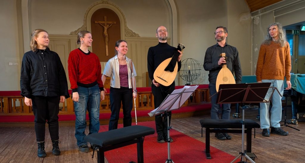 Kaikki kuusi esiintyjää seisomassa rivissä hymyille, osalla käsissään soittimia.